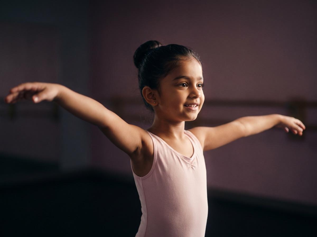 A smiling young girl in a pink ballet leotard practises with her arms outstretched in a softly lit dance studio